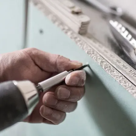 Eine Hand benutzt eine Bohrmaschine an einer Wand, mit einem Lineal und anderen Werkzeugen in der Nähe.