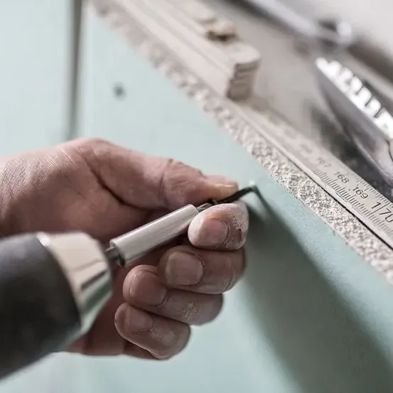 Eine Hand benutzt eine Bohrmaschine an einer Wand, mit einem Lineal und anderen Werkzeugen in der Nähe.