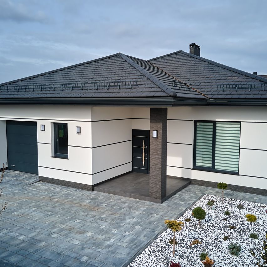 Ein modernes weißes Haus mit grauem Dach und Einfahrt. Das Haus hat eine Garage und eine Veranda mit zwei Säulen. Der Vorgarten hat einen kleinen Garten mit weißen Kieselsteinen.