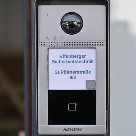 Ein Nahaufnahme eines Hikvision-Videotürgongs mit einer Sicherheitskamera, einem Mikrofonsymbol und einem kleinen Bildschirm, auf dem die Adresse 'Effenberger Sicherheitstechnik, St. Pöltnerstraße 8/2' steht.