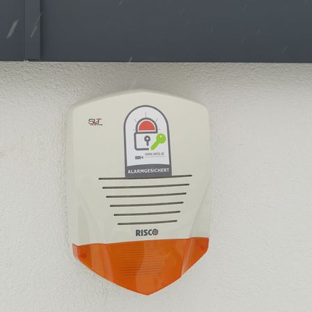 Ein weißer und oranger Alarmanlage mit der Aufschrift 'RISCO' ist an einer weißen Wand befestigt. Die Alarmanlage hat ein Schlosssymbol und den Text 'ALARMGESICHERT'.