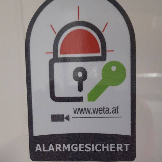 Das Bild zeigt einen Aufkleber für Alarmschutz. Es zeigt ein Schlosssymbol mit einem Schlüssel daneben und ein rotes Alarmsymbol. Die Website www.weta.at ist ebenfalls angezeigt. Der Text 'ALARMGESICHERT' befindet sich unter dem Symbol.