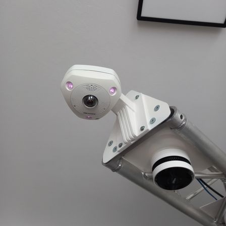 Eine weiße Kamera mit lila Lichtern ist an einer weißen Wand an einer Metallstruktur befestigt. Ein schwarzer Rahmen ist ebenfalls an der Wand befestigt.