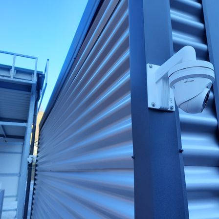 Eine weiße Überwachungskamera ist an einer blauen Metallwand mit Wellblech angebracht. Eine blaue Metallbrüstung befindet sich auf der linken Seite.