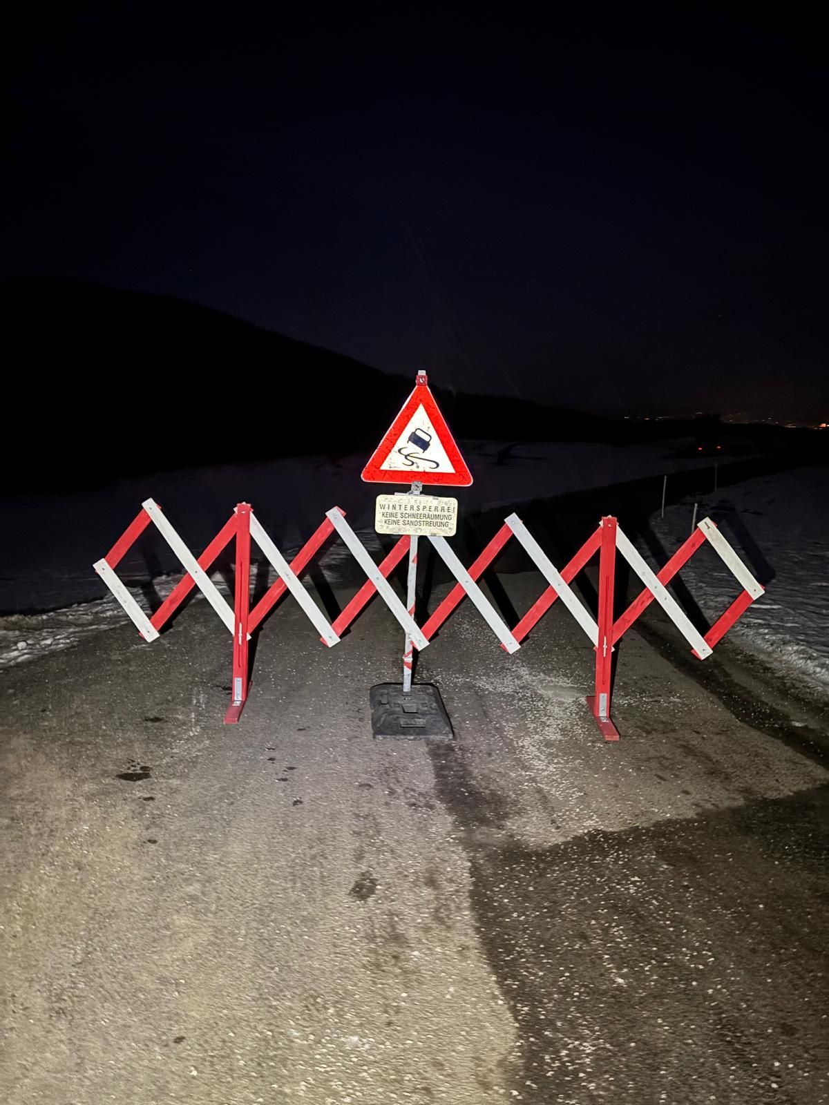 Ein Straßenbarriere mit einem dreieckigen Warnschild steht auf einer verschneiten Straße bei Nacht. Das Schild sagt 'WINTERSPERR' und darunter 'KEINE SCHNEEBESCHIEUNG'.