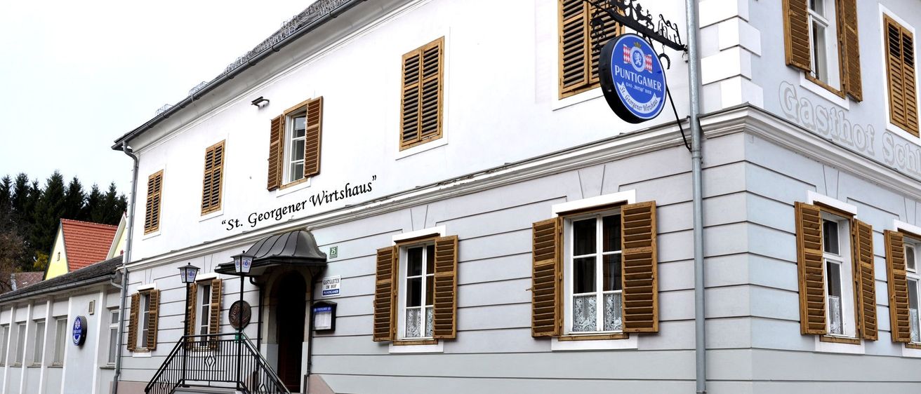 Ein weißes Gebäude mit braunen Fensterläden und einem blauen Schild mit der Aufschrift "Puntigamer". Es hat eine Treppe mit schwarzer Brüstung, die zum Eingang führt. Das Schild "St. Georgener Wirtshaus" ist an der Wand angebracht.