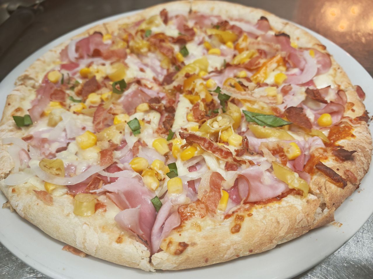 Eine Pizza mit Schinken, Mais, Käse und Speck auf einem weißen Teller.