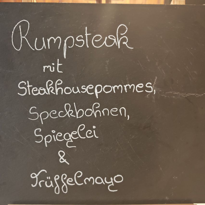 Ein schwarzes Menu-Brett zeigt Rumpsteak mit Steakhousepommes, Speckbohnen, Spiegelei und Truffelmayo an.