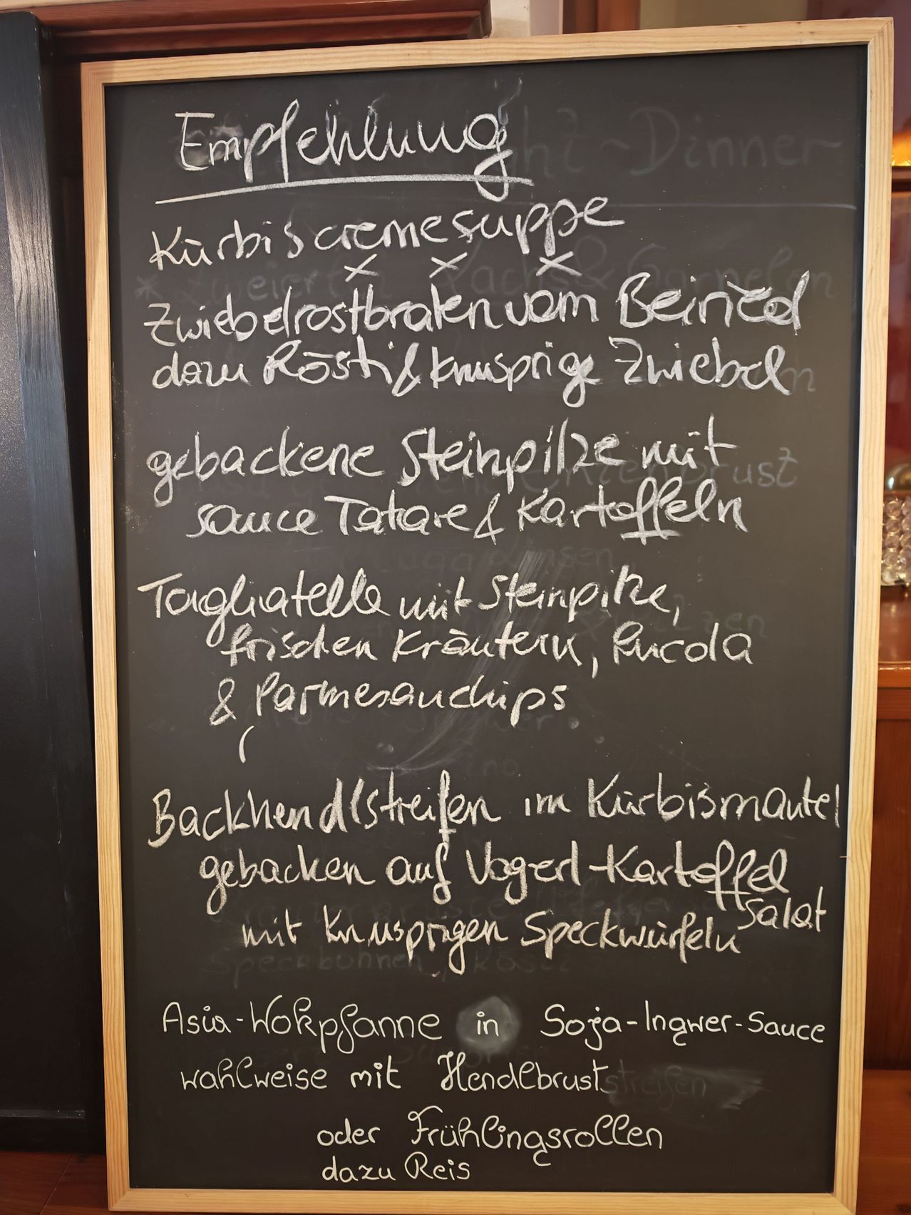 Ein Tafel zeigt eine Speisekarte mit Gerichten wie 'Curry-Cremesuppe', 'Gebratener Rinderbraten', 'Gegrilltes Steak' und 'Spargel in Soja-Ingwer-Sauce'.