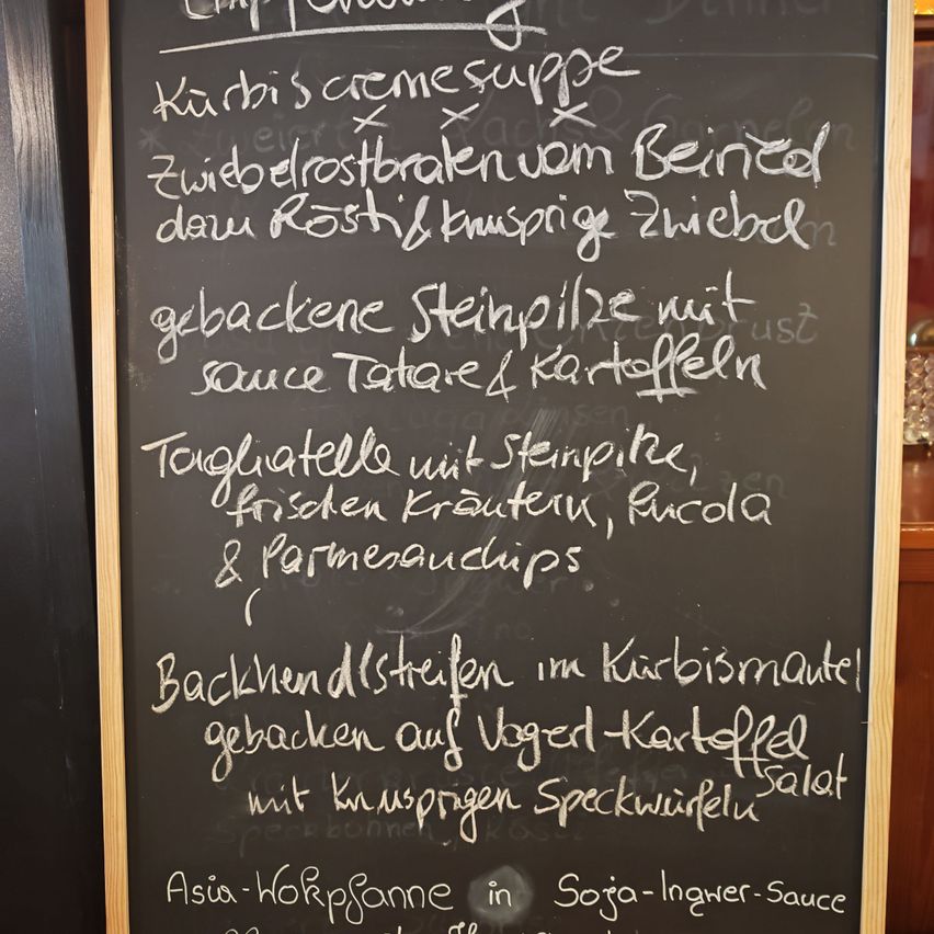 Ein Tafel zeigt eine Speisekarte mit Gerichten wie 'Curry-Cremesuppe', 'Gebratener Rinderbraten', 'Gegrilltes Steak' und 'Spargel in Soja-Ingwer-Sauce'.