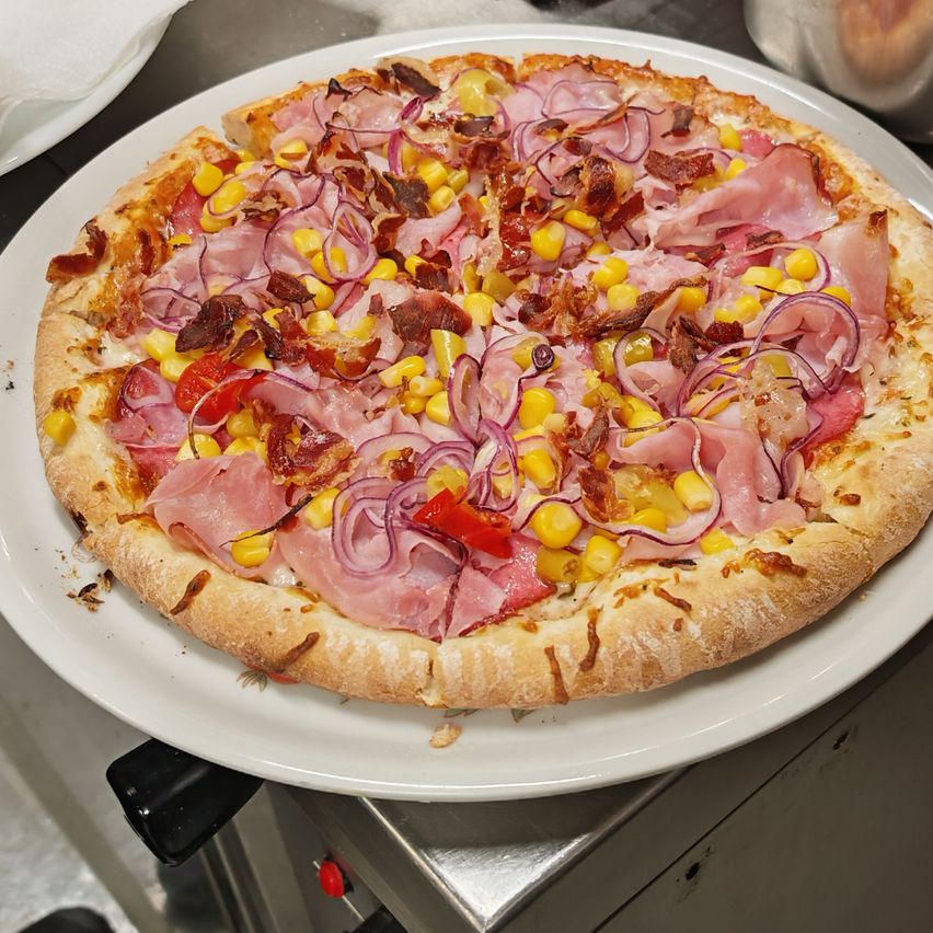 Eine Pizza mit Mais, roten Zwiebeln, Speck und Schinken steht auf einem weißen Teller in der Küche.