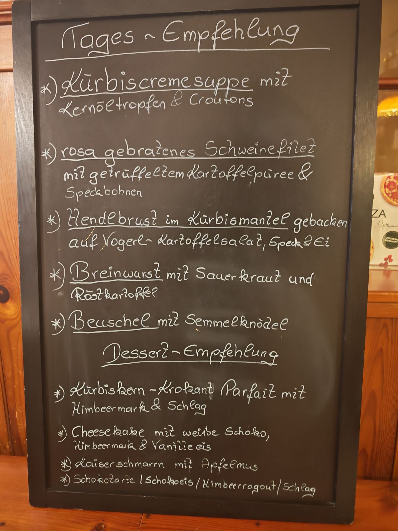 Ein Kreidetafel listet ein Menü mit Kürbiscremesuppe, gebratenem Schweinefilet, Hähnchenbrust, Bratwurst und Dessertempfehlungen auf. Gerichte umfassen Kürbiskerne, Käsekuchen und Kaiserschmarrn.
