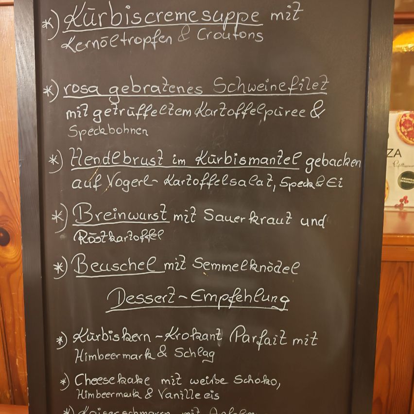 Ein Kreidetafel listet ein Menü mit Kürbiscremesuppe, gebratenem Schweinefilet, Hähnchenbrust, Bratwurst und Dessertempfehlungen auf. Gerichte umfassen Kürbiskerne, Käsekuchen und Kaiserschmarrn.