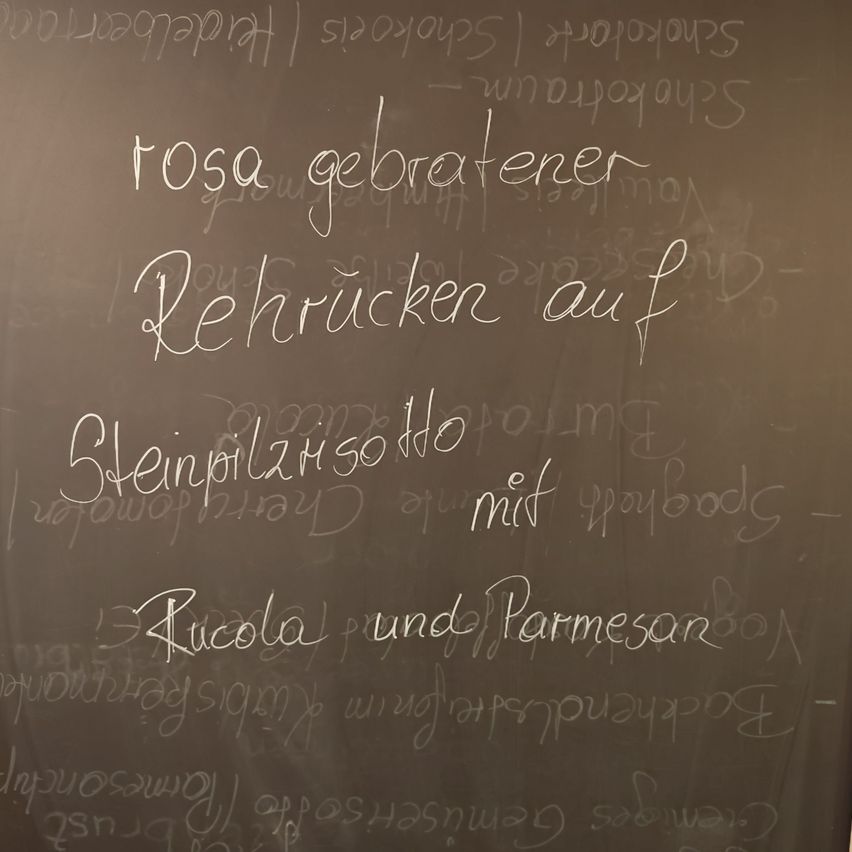 Eine Kreidetafel zeigt handgeschriebenen Text in weißer Kreide. Der Text enthält Wörter wie 'Rosa', 'Rehrücken', 'Steinpilzsoße' und 'Rucola und Parmesan'. Es scheint eine Liste oder ein Rezept zu sein.