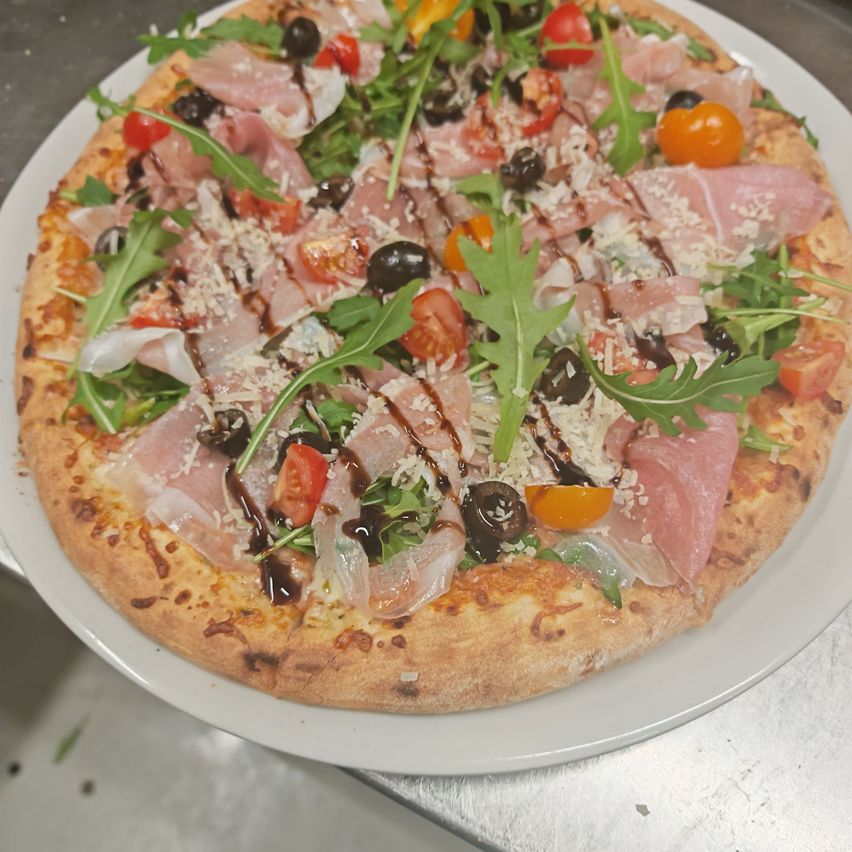 Eine runde Pizza, belegt mit Käse, Kirschtomaten, schwarzen Oliven, Rucola und Prosciutto-Scheiben, garniert mit brauner Sauce und serviert auf einem weißen Teller auf einer Metalloberfläche.