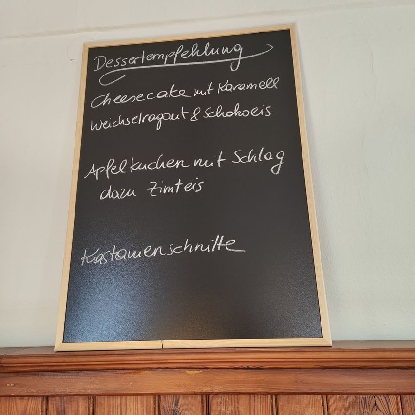 Eine Tafel zeigt Dessertempfehlungen, darunter Cheesecake mit Karamell, wechselndes Dessert und Apfelkuchen mit Schlagsahne, gekrönt mit Zimt.