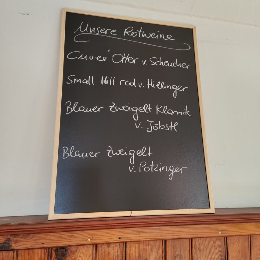 Eine Tafel zeigt verschiedene Rotweine von verschiedenen Winzern. Oben steht 'Unsere Rotweine', gefolgt von Namen der Weine und Winzer. Darunter steht 'Blauer Zweigelt' und 'v. Potzinger'. Die Tafel befindet sich auf einer Holzoberfläche.