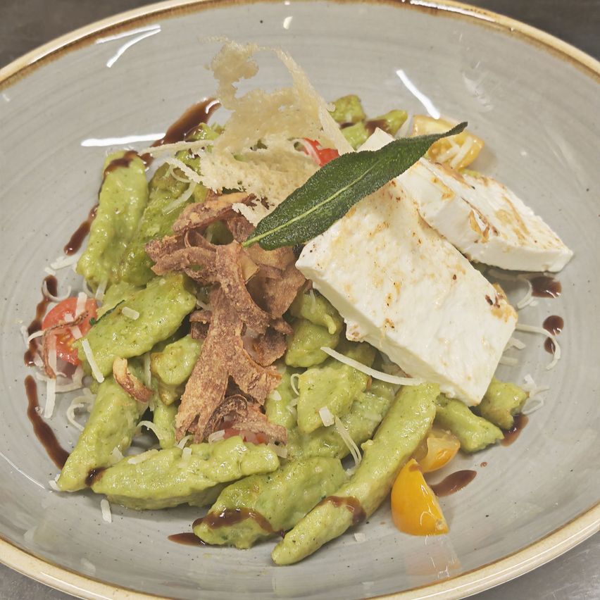 Ein Teller mit grüner Pasta in Avocado-Sauce, garniert mit Feta, Cherry-Tomaten und knusprigen Speckstückchen.