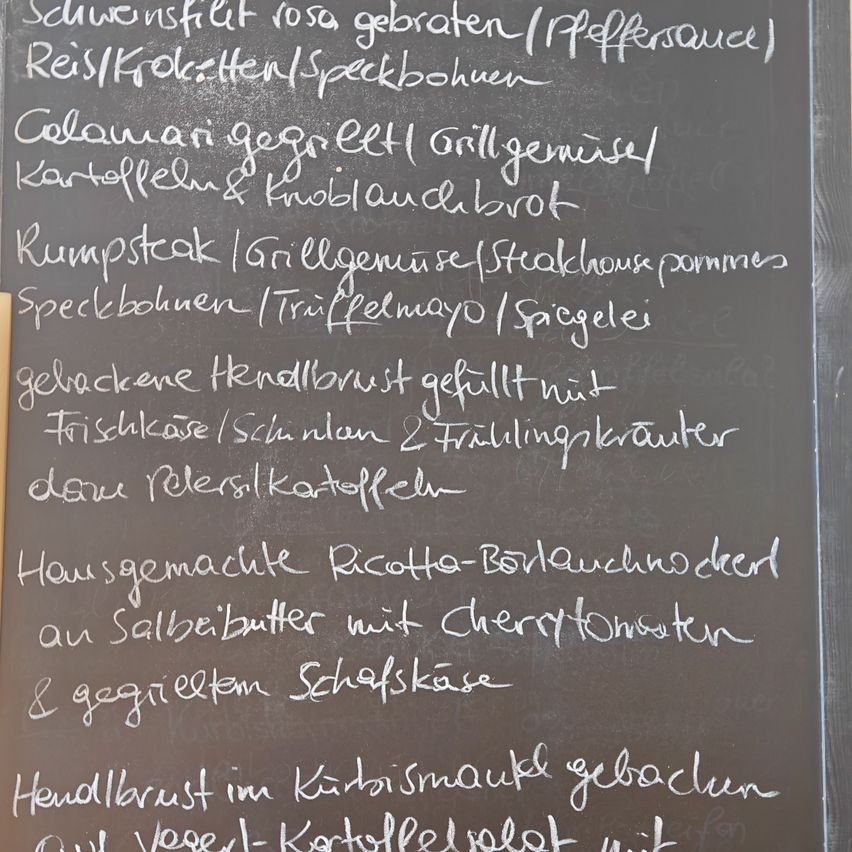 Eine Tafel listet verschiedene Gerichte auf, darunter geräuchertes Schweinefleisch, gegrilltes Gemüse und hausgemachte Ricotta mit Kirschtomaten und Ziegenkäse.