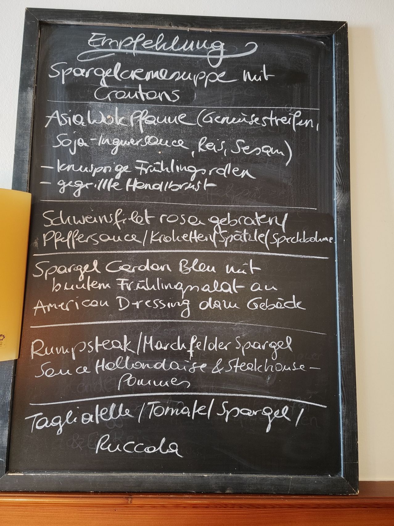 Eine Kreidetafel zeigt eine Speisekarte mit mehreren Essensoptionen, die sowohl auf Englisch als auch auf Deutsch geschrieben sind. Zuerst gibt es ein Gericht mit Croutons, gefolgt von einem asiatischen Gericht mit Sojasauce. Die Speisekarte enthält auch eine Vielzahl anderer Gerichte wie ein Steak mit Spargel und Saucen.