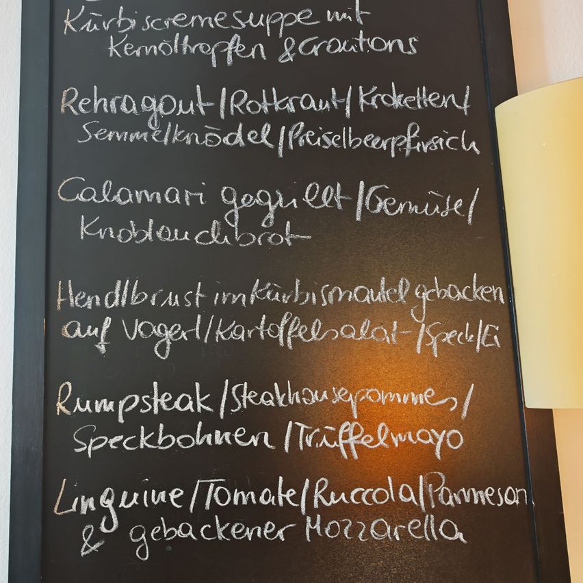 Ein Kreidetafel zeigt ein Menü mit verschiedenen Gerichten wie Rehaglut, Calamari, Rumpsteak und Linguine. Jedes Gericht ist mit seinen Zutaten oder Zubereitungsart aufgeführt.