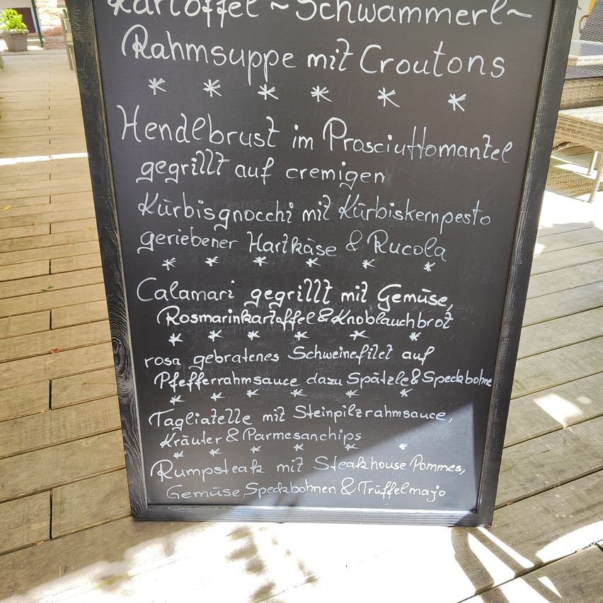 Ein Kreidetafel-Menü mit verschiedenen Gerichten, darunter 'Kartoffel Schwammerl Rahmsuppe mit Croutons' und 'Calamari gegrillt mit Gemuse'. Das Menü ist in weißer Kreide auf schwarzem Hintergrund geschrieben.