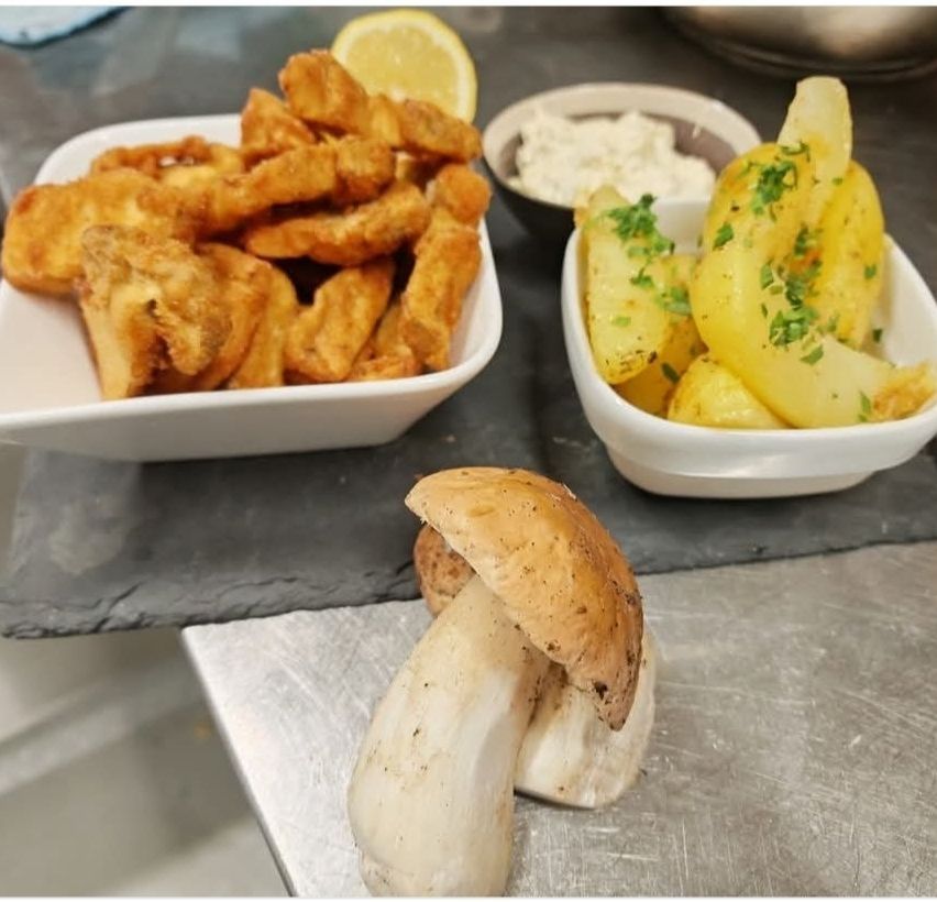 Ein Tisch mit zwei weißen Schüsseln, eine mit frittierten Fischstücken und die andere mit Kartoffelscheiben, die mit Kräutern garniert sind, zusammen mit einem Pilz auf einer Metalloberfläche.