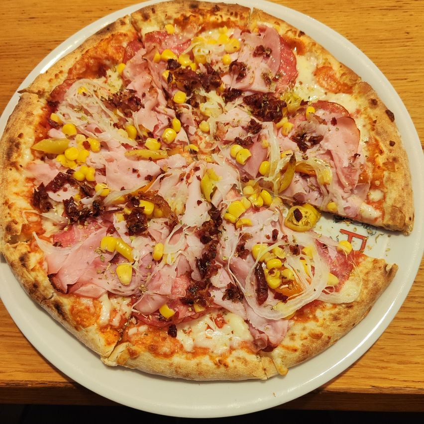 Eine runde Pizza mit Schinken, Mais, Zwiebeln und Käse auf einem weißen Teller auf einem Holztisch.