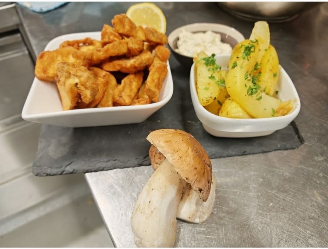 Ein Teller mit frittierten Fischfingern, eine Schüssel mit Kartoffelspalten mit Kräutern und ein Pilz auf einer Metalloberfläche.