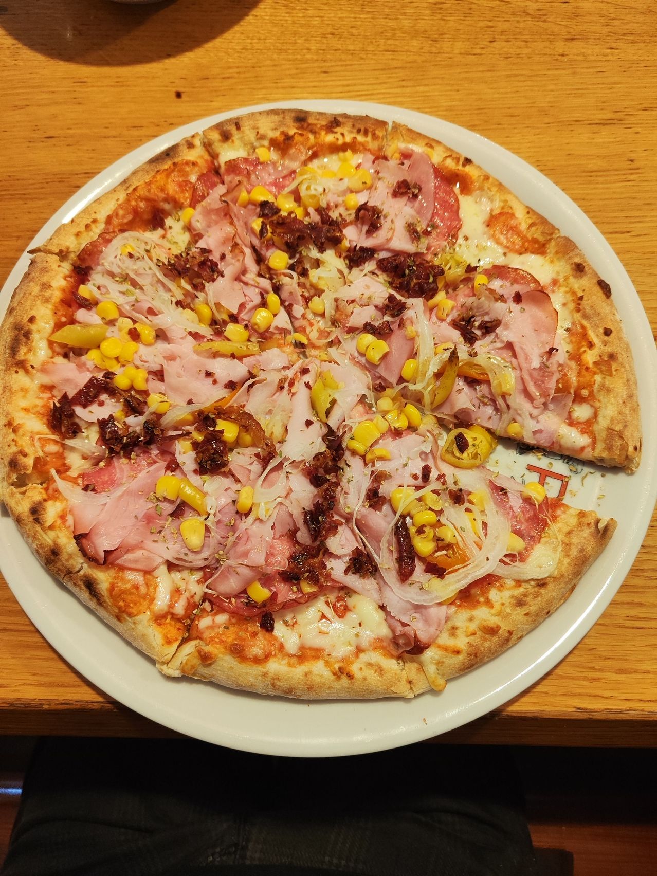 Eine runde Pizza mit Schinken, Mais, Zwiebeln und Käse auf einem weißen Teller auf einem Holztisch.