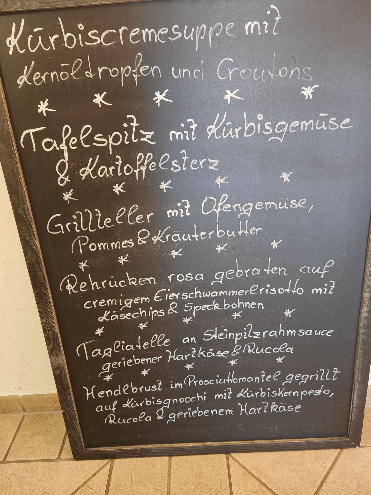 Ein Kreidetafel-Menü mit verschiedenen Gerichten wie 'Kernol Dropfen und Croutons', 'Tafelspitze mit Kurbisgemüse & Kartoffelsterz', 'Grillteller mit Ofengemüse, Pommes & Krauterbutter' und mehr.