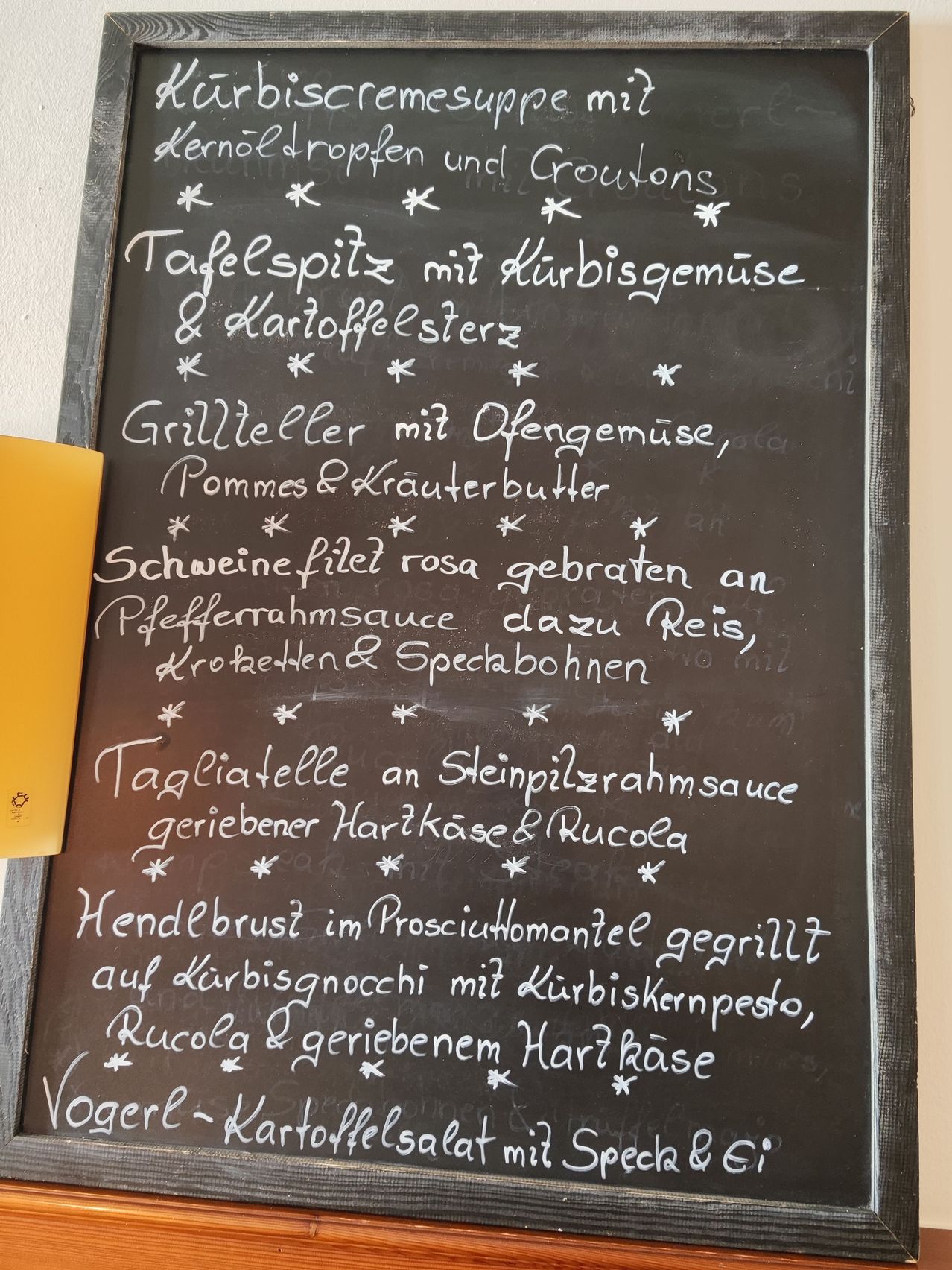 Ein Kreidetafel-Menü mit verschiedenen Gerichten, darunter 'Grillteller mit Ofengemüse,' 'Schweinefilet rosa gebraten an Pfefferrahmsauce' und 'Tagliatelle an Steinpilzrahmsauce.' Jedes Gericht ist mit einem Stern markiert.