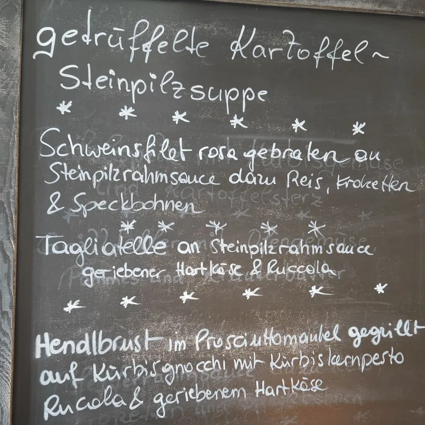 Ein Kreidetafelmenü mit deutschem Text. Es listet mehrere Gerichte auf, darunter getrüffelte Kartoffeln, Steinpilzsuppe und ein Gericht mit Schweinefleisch und Rahmsauce.