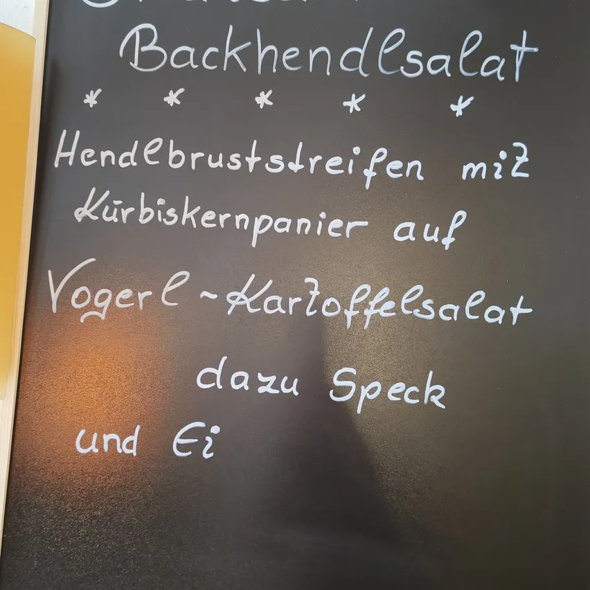 Ein Kreidetafel mit einem in weißer Kreide geschriebenen Menü. Das Menü umfasst Steirischer Backhendlsalat, ein Hühnchenbrustgericht mit Kürbiskernen und Ei.