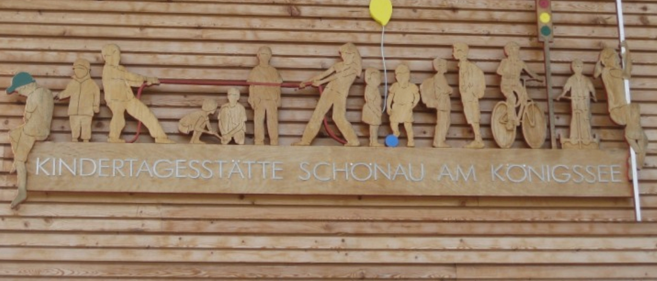 Ein Holzschild an einer Holzwand zeigt Figuren von Kindern und einem Ballon sowie den Text 'Kindertagesstätte Schonau am Konig'.