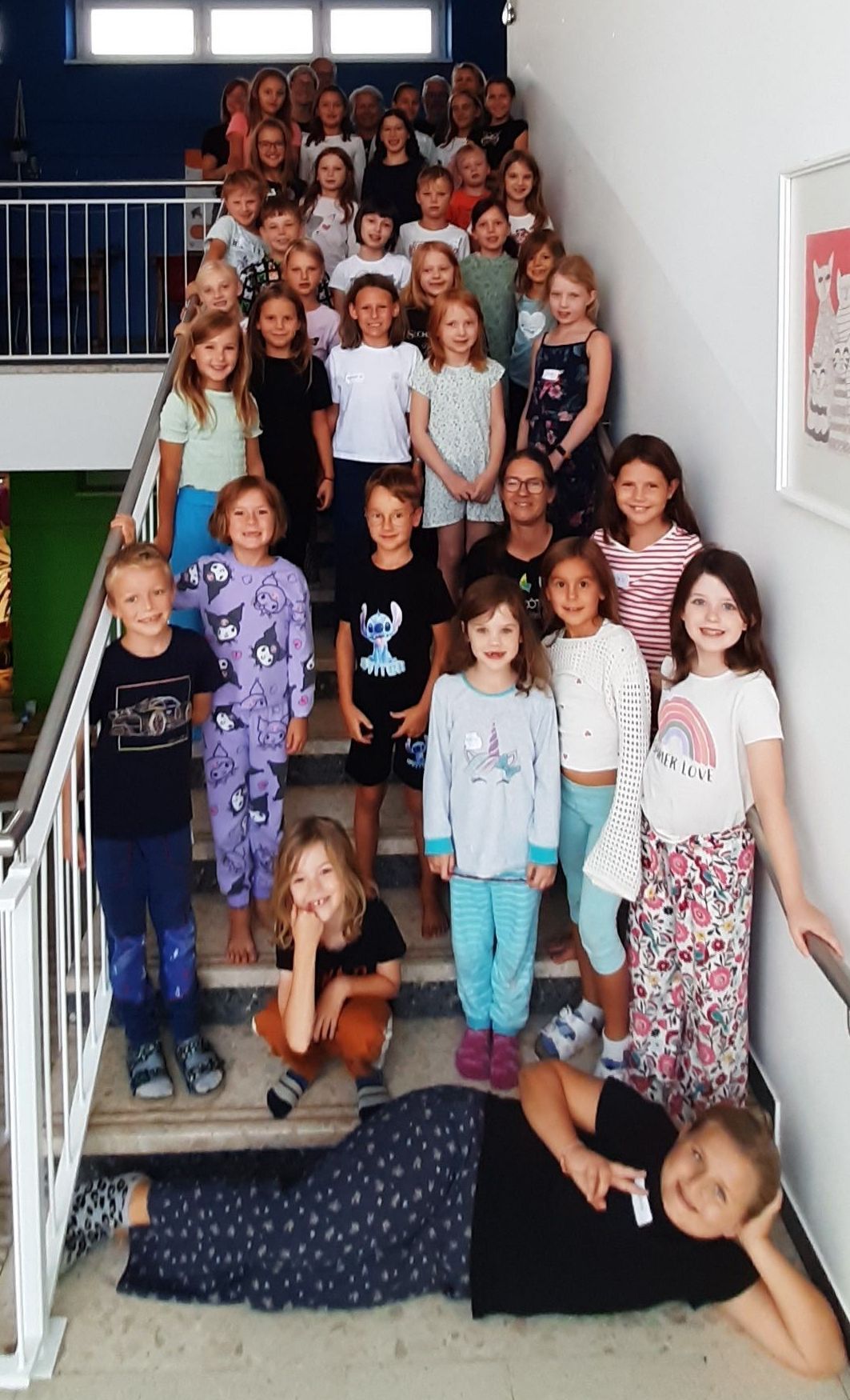 Eine Gruppe von Kindern und Erwachsenen steht auf den Stufen und lächelt für ein Foto. Einige tragen Schlafanzüge, andere Freizeitkleidung. Die Umgebung ist drinnen, wahrscheinlich eine Schule oder ein Gemeindezentrum.