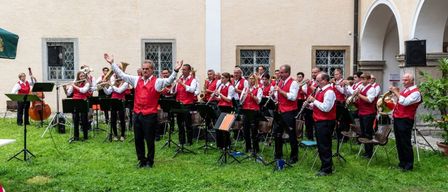 Eine Band spielt auf einem Rasenfeld vor einem Gebäude. Sie tragen rote Westen und schwarze Hosen. Der Dirigent leitet die Band.