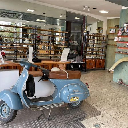 Ein altes blaues Vespa-Scooter steht in einem Geschäft mit gefliesten Böden und Holzregalen voller Brillen. Rechts sind ein roter Roller und ein Reifen.