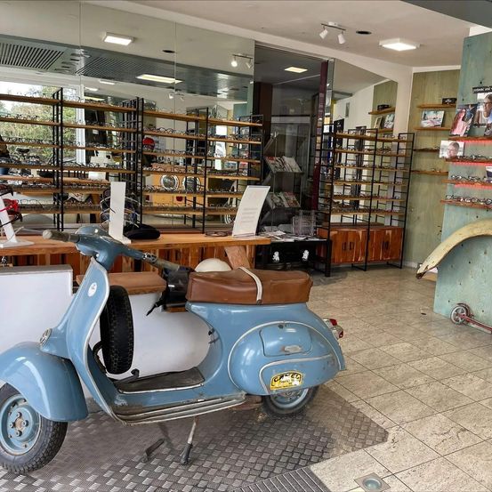 Ein altes blaues Vespa-Scooter steht in einem Geschäft mit gefliesten Böden und Holzregalen voller Brillen. Rechts sind ein roter Roller und ein Reifen.