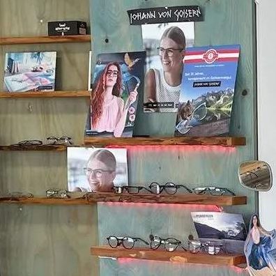 Ein Innenansicht eines Optikers mit einer Ausstellung verschiedener Brillen auf Regalen und einem großen Plakat, das 'UnerreA3LEED' bewirbt. Ein Vintage-stilvoller Wagen mit einem Reifen und einem Surfbrett steht in der Mitte.
