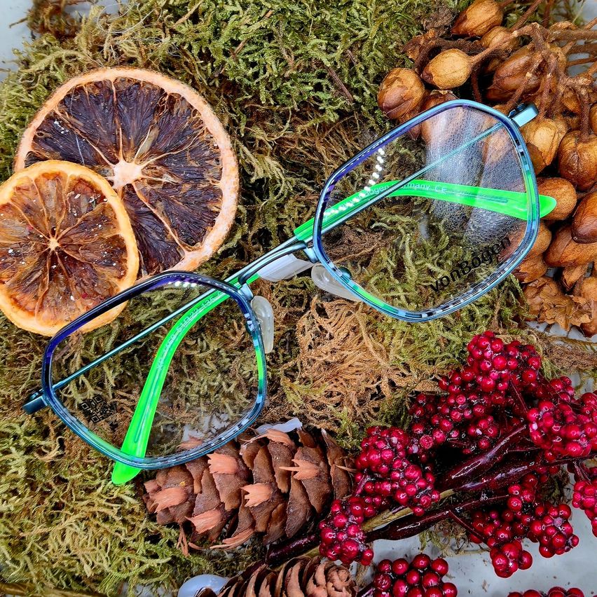 Ein Paar Brille mit grünen Rahmen liegt auf einem Moosbett, getrockneten Zitrusscheiben, Tannenzapfen und roten Beeren.