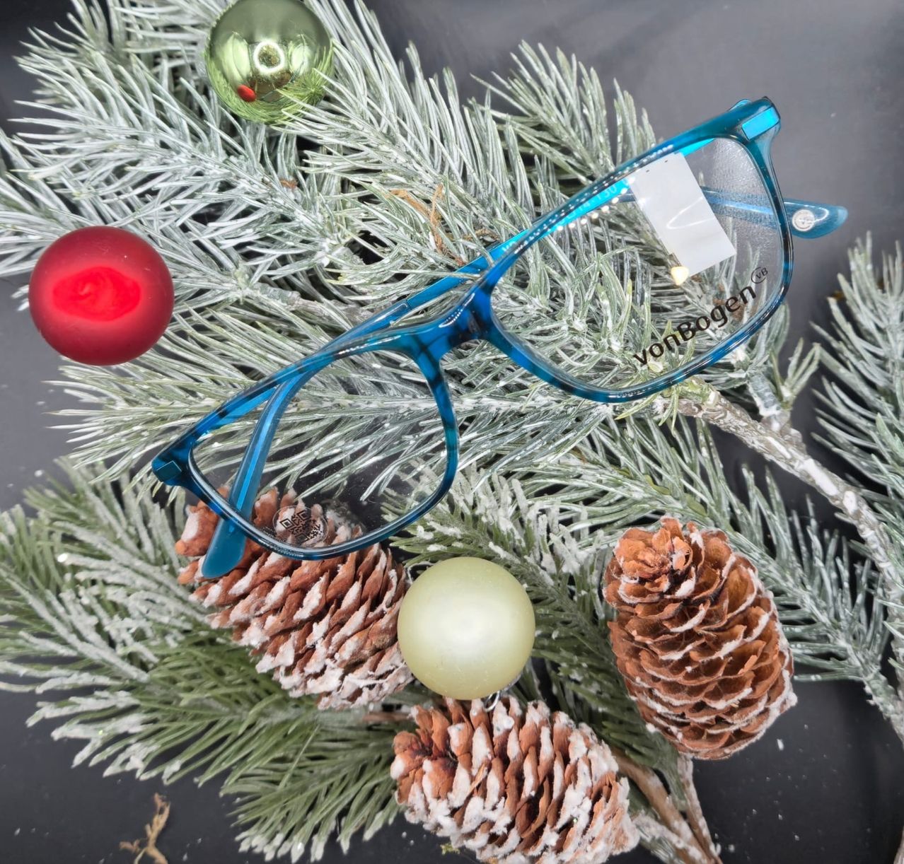 Ein Paar blaue Brille liegt auf einem Weihnachtsbaum, der mit Tannenzapfen und Ornamenten geschmückt ist. Die Brille ist mit 'vonBogen' beschriftet.