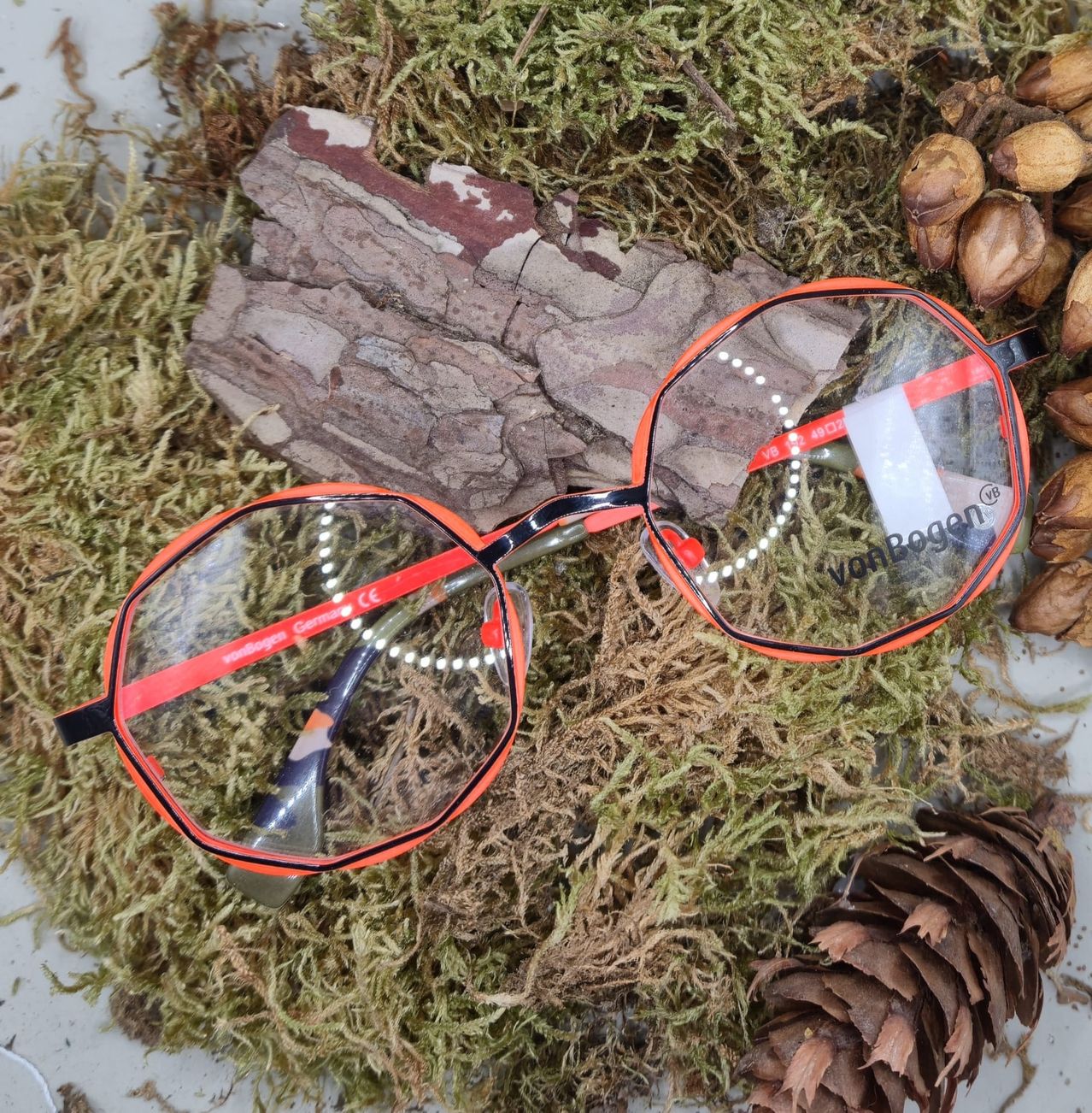 A pair of orange-framed glasses with clear lenses rests on a bed of moss, surrounded by pine cones and small stones.