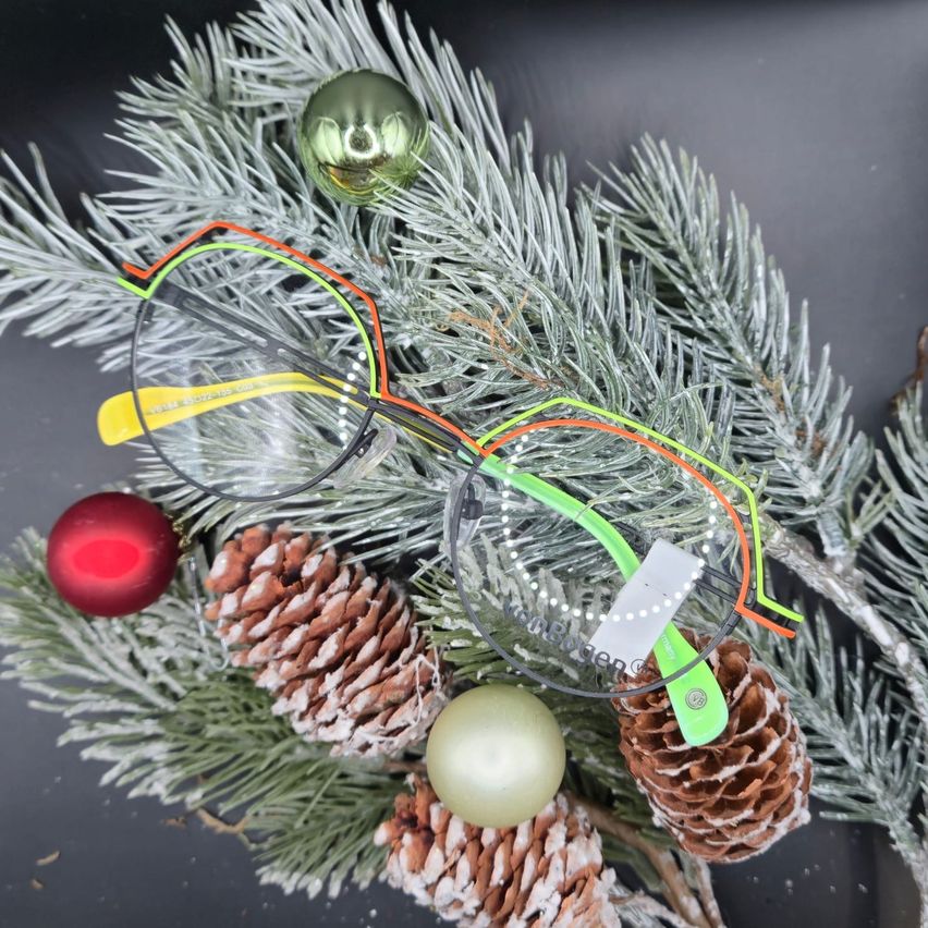 A pair of glasses with colorful frames rests on a decorated Christmas tree. The tree is adorned with pine cones, Christmas balls, and a green ornament. The background is black.