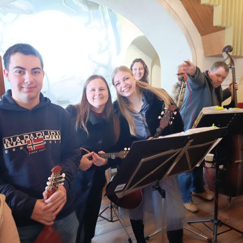 Vier Personen lächeln in einem Raum und halten Gitarren. Sie stehen vor einem Notenständer. Ein Mann rechts hält ein Cello. Die Frau in der Mitte hat lange blonde Haare.