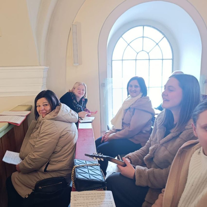 Fünf Frauen sitzen lächelnd in einer Kirche. Eine hält eine Gitarre, andere haben Notenblätter. Der Raum hat ein Bogenfenster und eine weiße Wand.