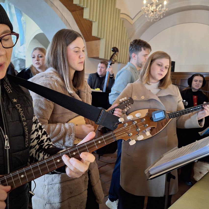 Eine Gruppe von Musikern spielt Saiteninstrumente in einer Innenumgebung. Zwei Frauen halten Gitarren, eine mit einem Gurt um die Schulter. Eine weitere Frau spielt Cello. Hinter ihnen befindet sich eine Treppe und eine Kristallleuchter.
