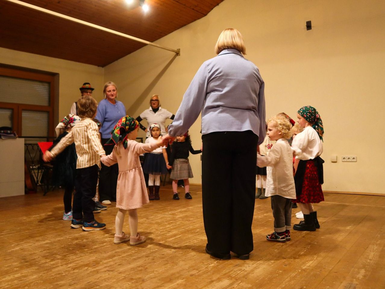 Eine Gruppe von Kindern und Erwachsenen tanzt in einem Raum mit Holzböden und weißen Wänden. Einige tragen traditionelle Kleidung.