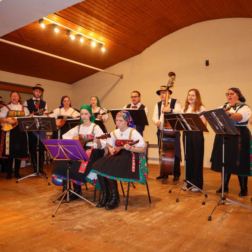Eine Gruppe von Musikern in traditionellen Kostümen spielt im Innenbereich. Sie spielen verschiedene Instrumente, darunter Gitarren und eine Geige. Der Raum hat Holzböden und eine weiße Wand.
