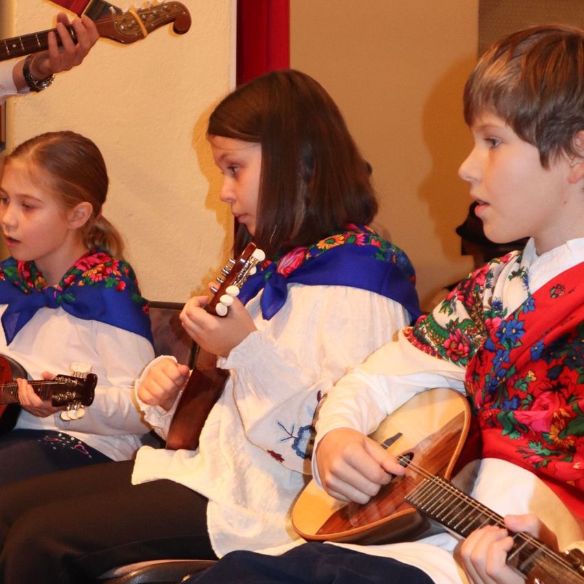 Drei Kinder sitzen und tragen traditionelle Kleidung, während sie Saiteninstrumente spielen. Das Kind auf der linken Seite spielt eine Gitarre, das in der Mitte eine Balalaika. Der Junge auf der rechten Seite spielt eine Mandoline.